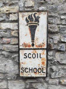 Weathered cast-iron Scoil / School sign on a stone wall, part of the Hands Exhibition at Sligo Folk Park